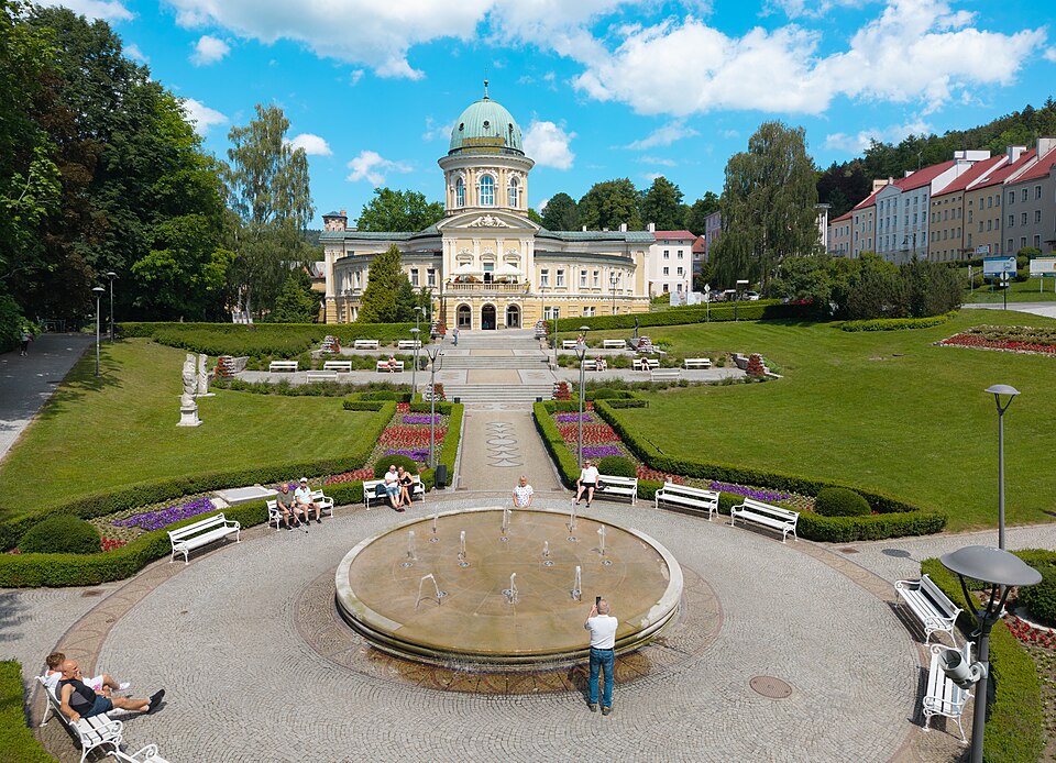 Fontanna, rabaty kwiatowe i zabytkowy budynek w Parku Zdrojowym w Lądku-Zdroju